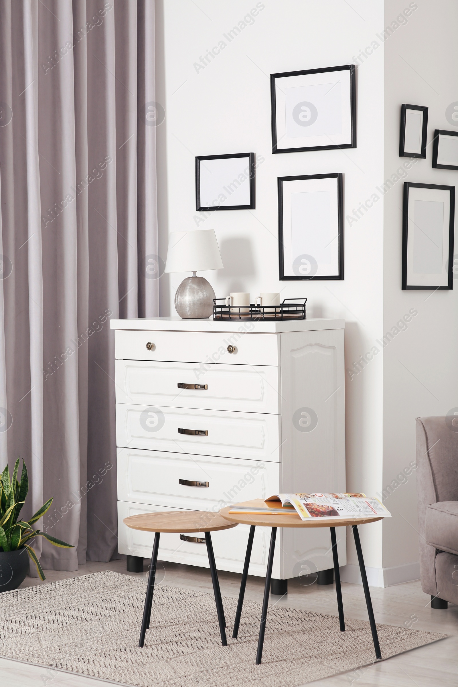 Photo of Empty frames hanging on white wall, chest of drawers and wooden tables indoors