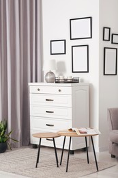 Empty frames hanging on white wall, chest of drawers and wooden tables indoors