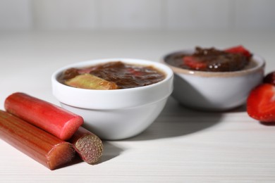 Photo of Tasty rhubarb jam in bowls, cut stems and strawberries on white wooden table