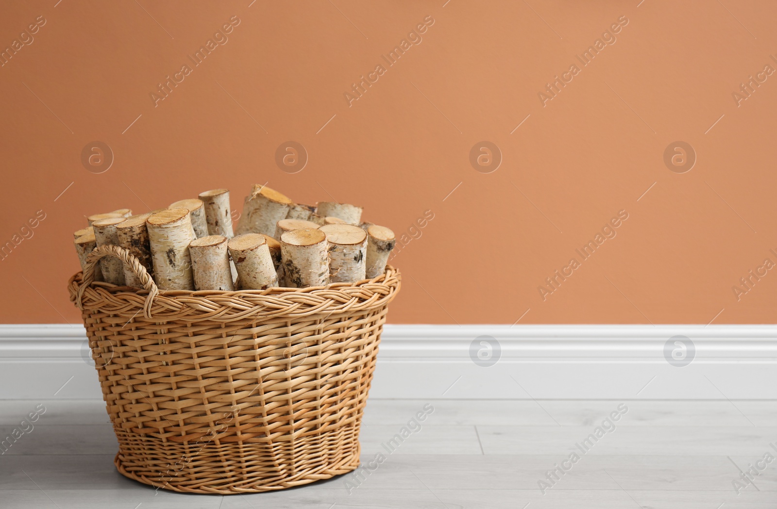 Photo of Wicker basket with firewood near brown wall indoors, space for text
