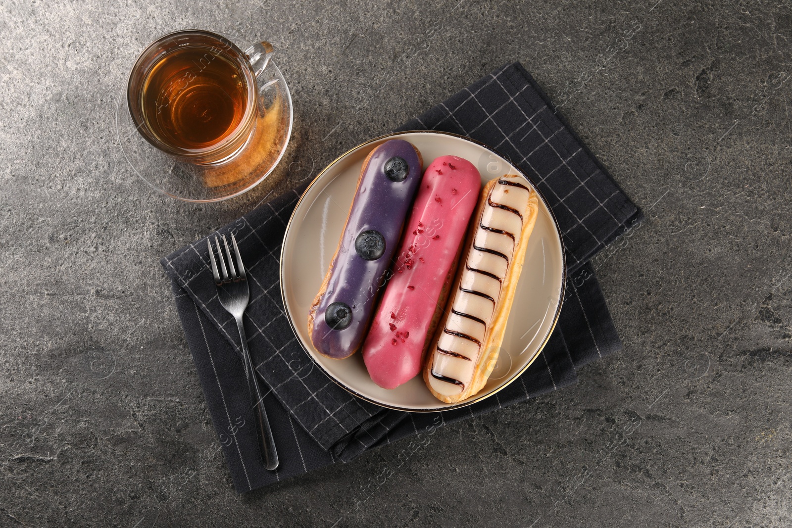 Photo of Different tasty glazed eclairs served with tea on grey textured table, flat lay