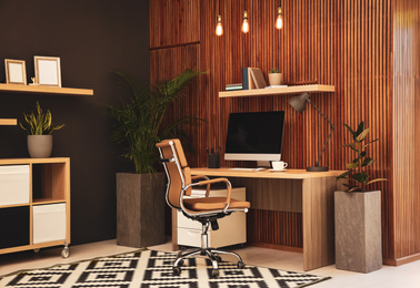 Photo of Comfortable workplace with computer near wooden wall in stylish room interior. Home office design