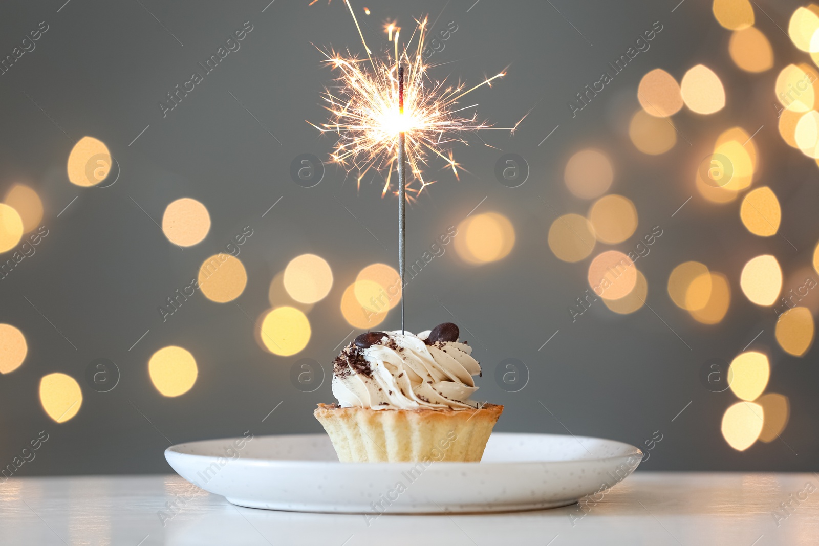 Photo of Cupcake with burning sparkler on table against blurred festive lights