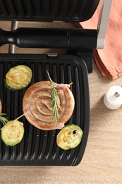 Photo of Electric grill with homemade sausage, zucchini and rosemary on wooden table, flat lay