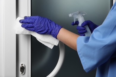 Photo of Woman in gloves cleaning door handle with paper towel and detergent indoors, closeup