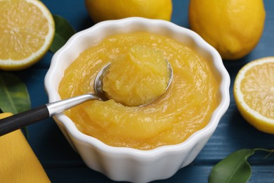 Photo of Delicious lemon curd in bowl, fresh citrus fruits and spoon on blue wooden table, closeup