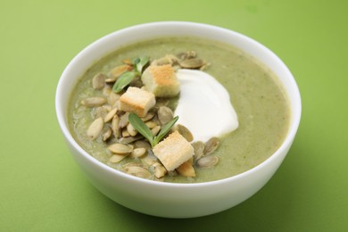 Photo of Delicious broccoli cream soup with croutons, sour cream and pumpkin seeds on green background, closeup