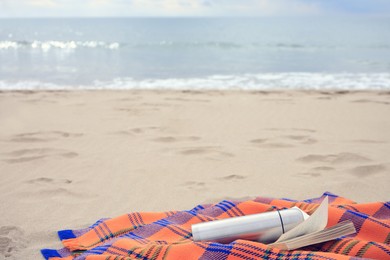 Metallic thermos with hot drink, open book and plaid on sandy beach near sea, space for text