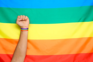 Man wearing gay wristband on hand near rainbow flag, closeup. Space for text