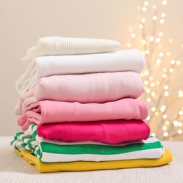 Photo of Stack of folded clothes on wooden table against beige background with blurred lights, space for text