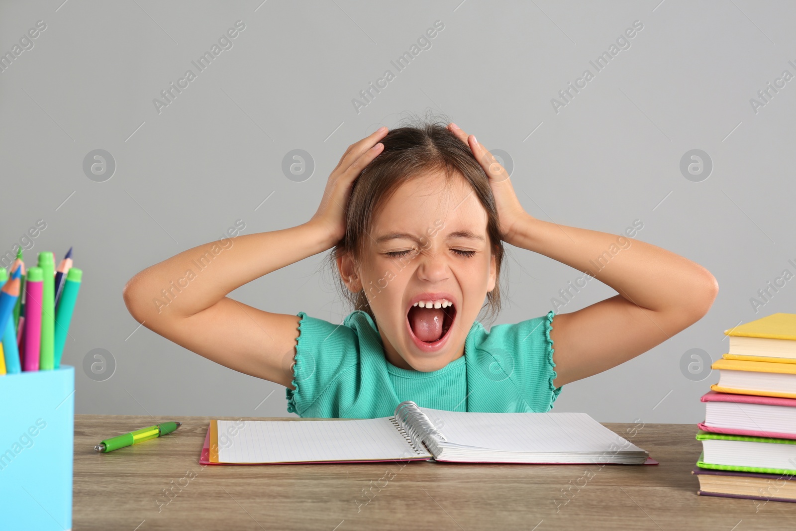 Photo of Emotional little girl doing homework at table on grey background