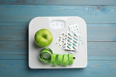 Scales with weight loss pills, apple and measuring tape on light blue wooden table, top view