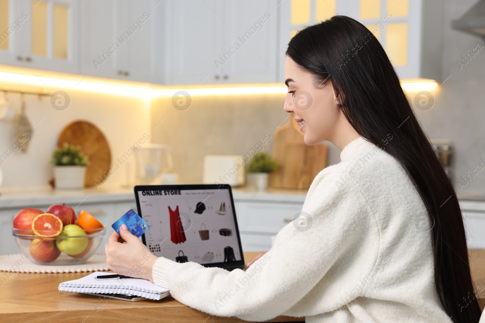 Photo of Young woman with credit card and laptop shopping online at wooden table in kitchen