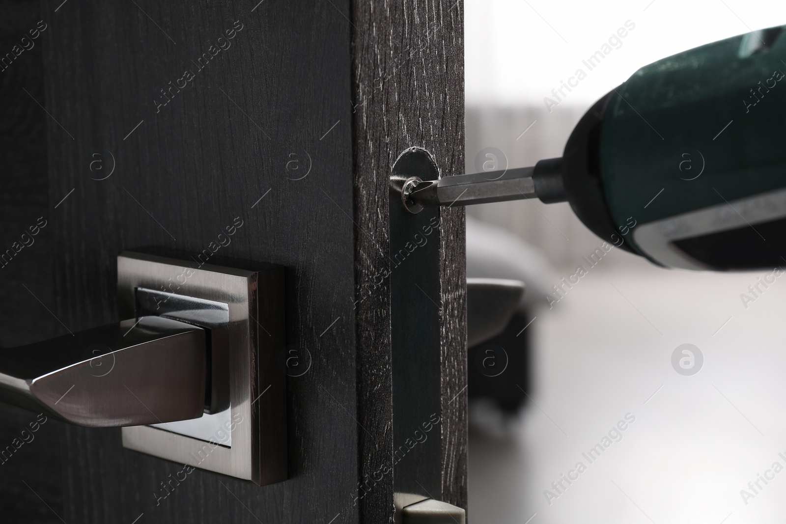 Photo of Handyman with electric screwdriver repairing door handle indoors, closeup