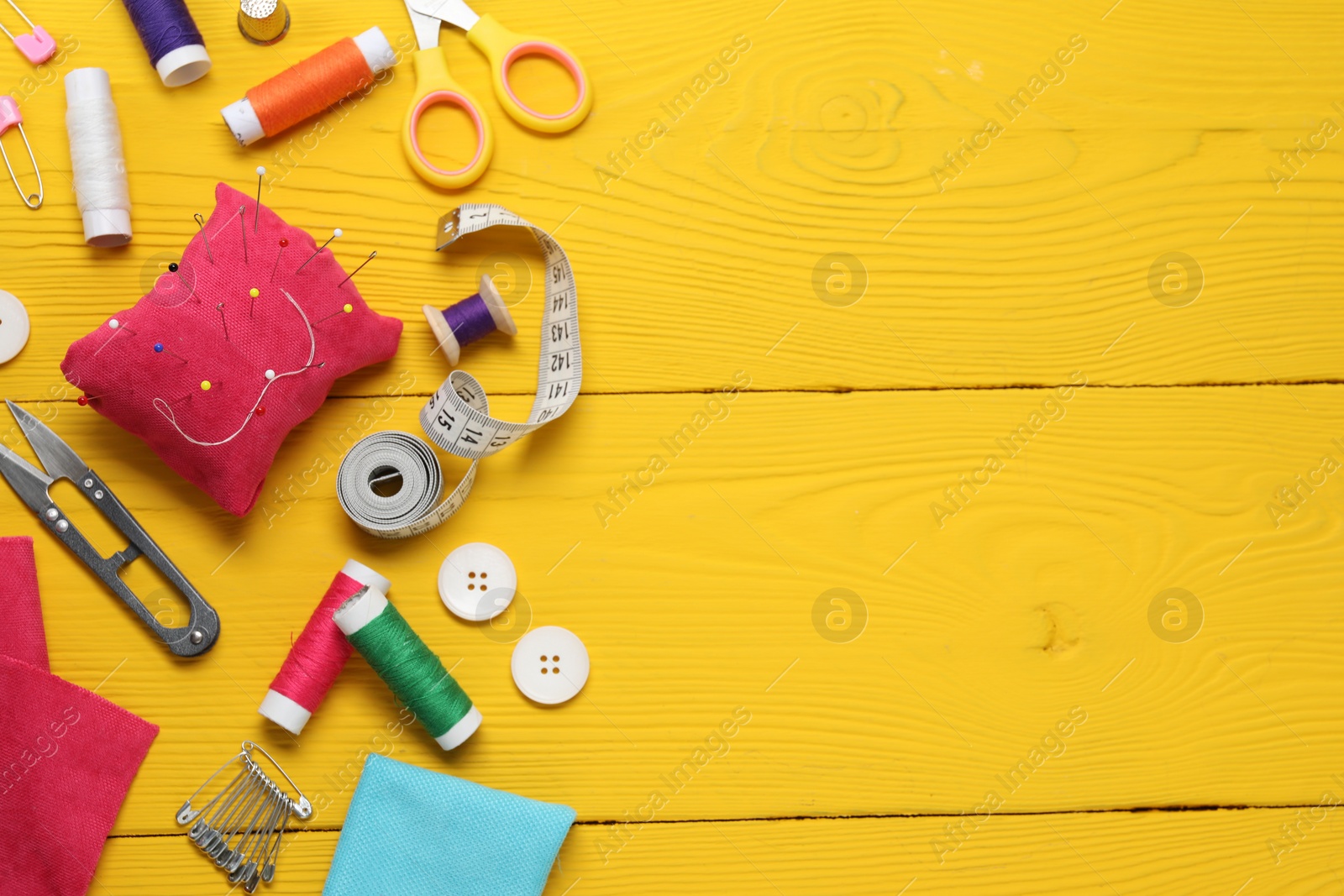 Photo of Red pincushion with pins and other sewing tools on yellow wooden table, flat lay. Space for text