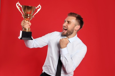 Photo of Portrait of happy young businessman with gold trophy cup on red background