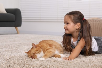 Photo of Smiling little girl petting cute ginger cat on carpet at home, space for text