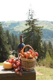 Photo of Basket for picnic with grapes, cheese and bottle of wine on wooden bench against mountain landscape