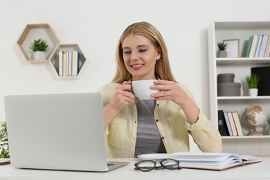 Photo of Home workplace. Happy woman with cup of hot drink looking at laptop at white desk in room