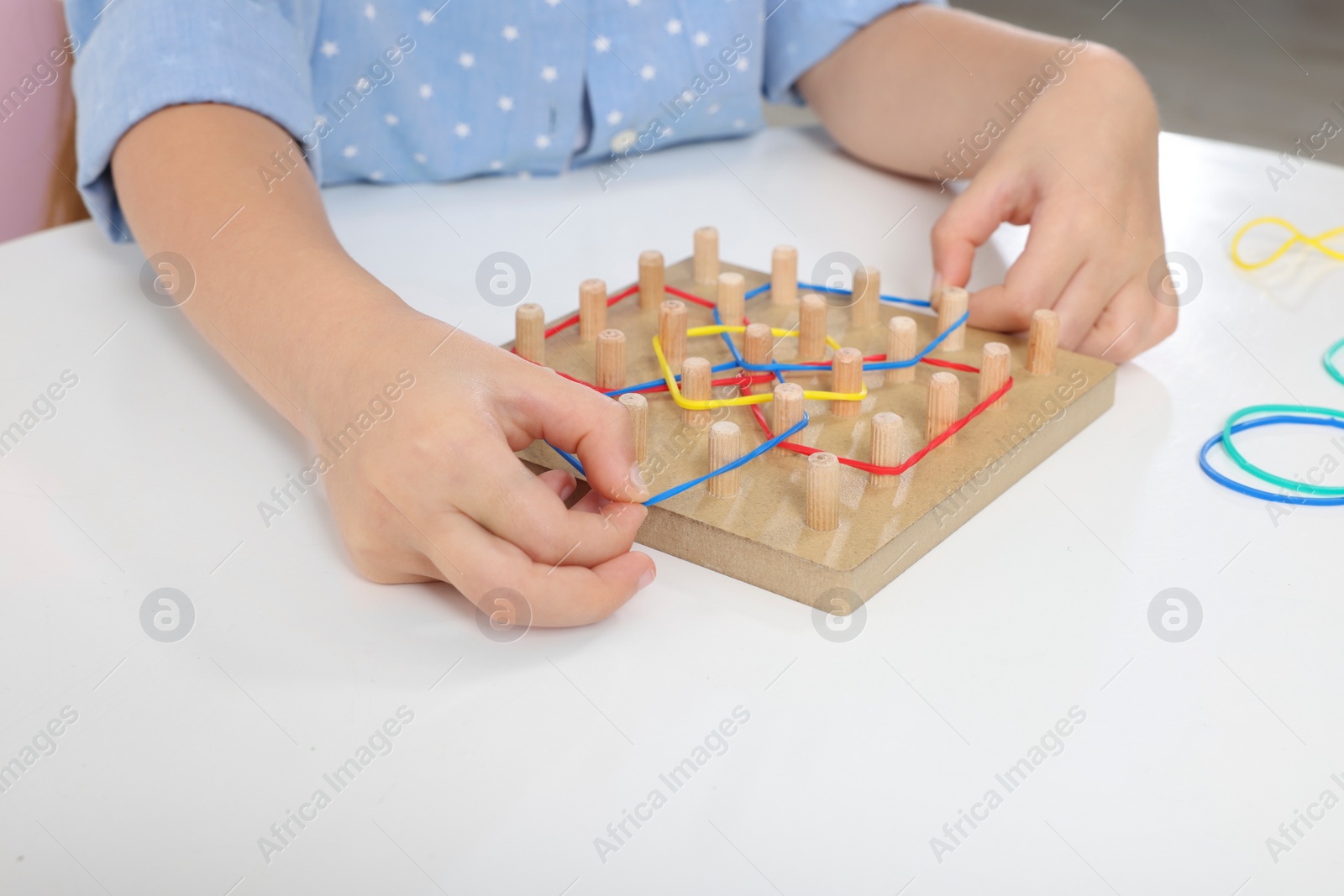 Photo of Motor skills development. Girl playing with geoboard and rubber bands at white table, closeup