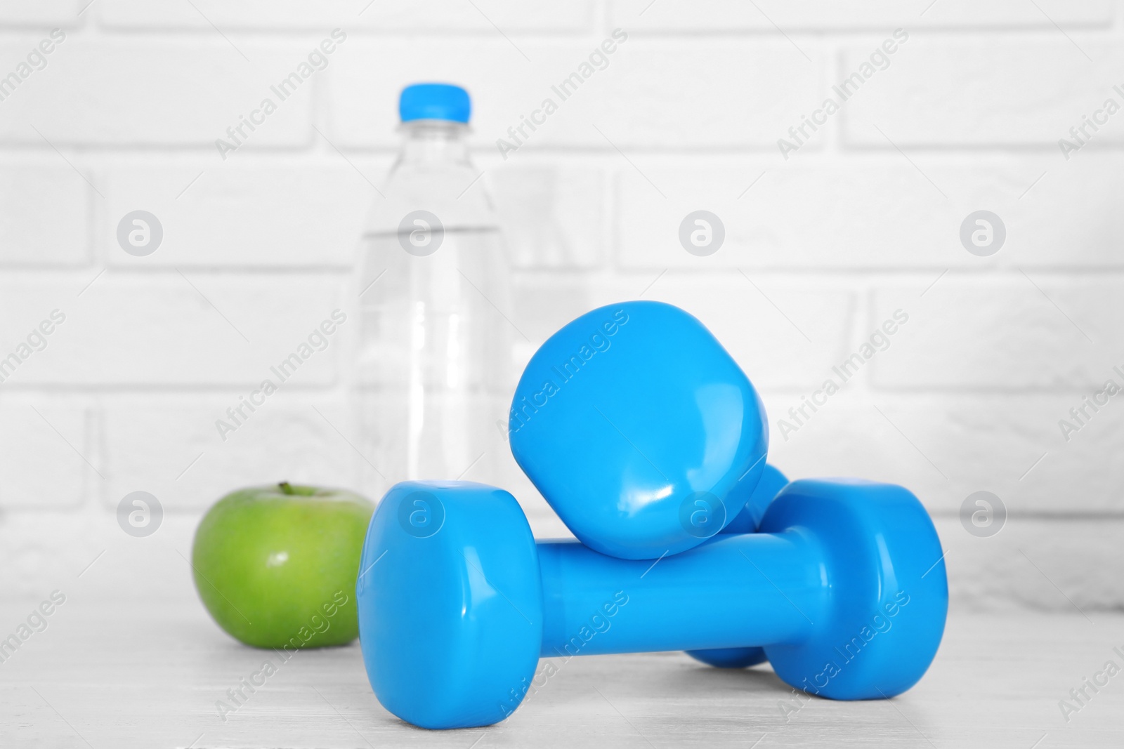 Photo of Stylish dumbbells, apple and bottle of water on table against brick wall. Home fitness