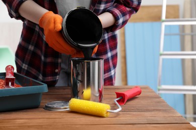 Photo of Woman pouring black paint from bucket into can at wooden table indoors, closeup