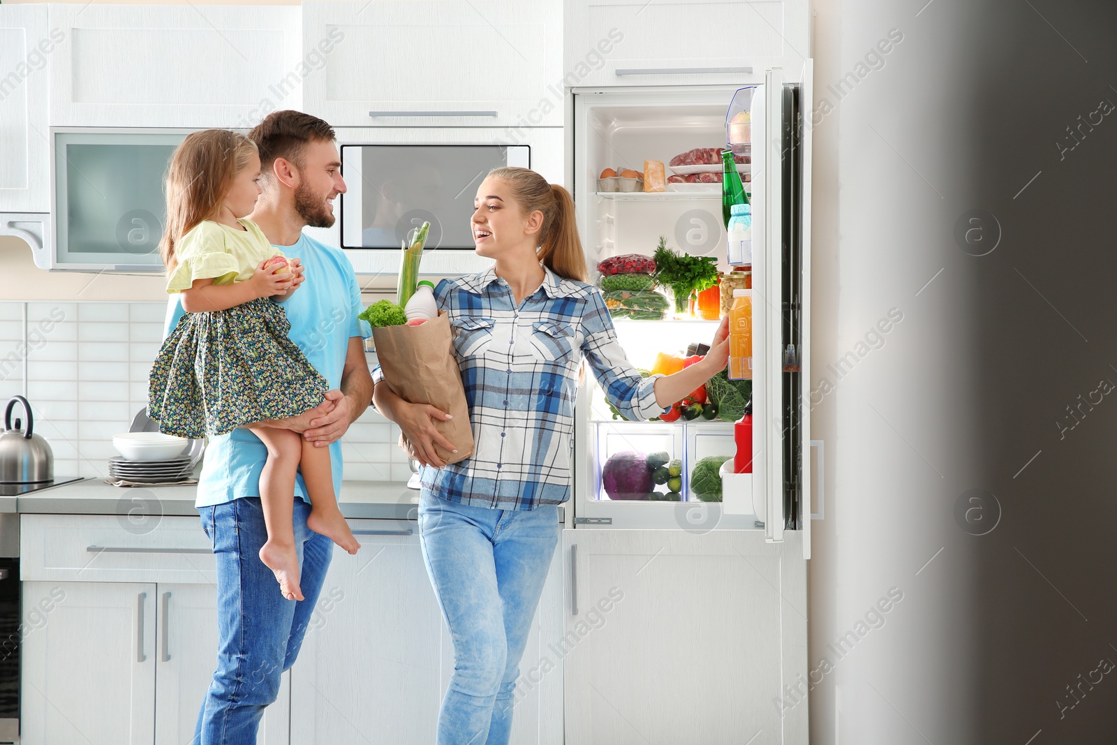 Photo of Happy family with paper bag full of products standing near refrigerator in kitchen