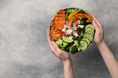 Photo of Woman holding bowl with many different vegetables and tofu at grey table, top view and space for text. Vegan diet