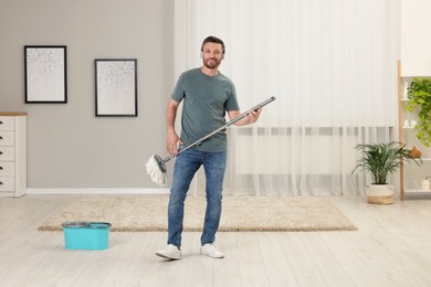 Photo of Enjoying cleaning. Happy man in headphones with mop at home