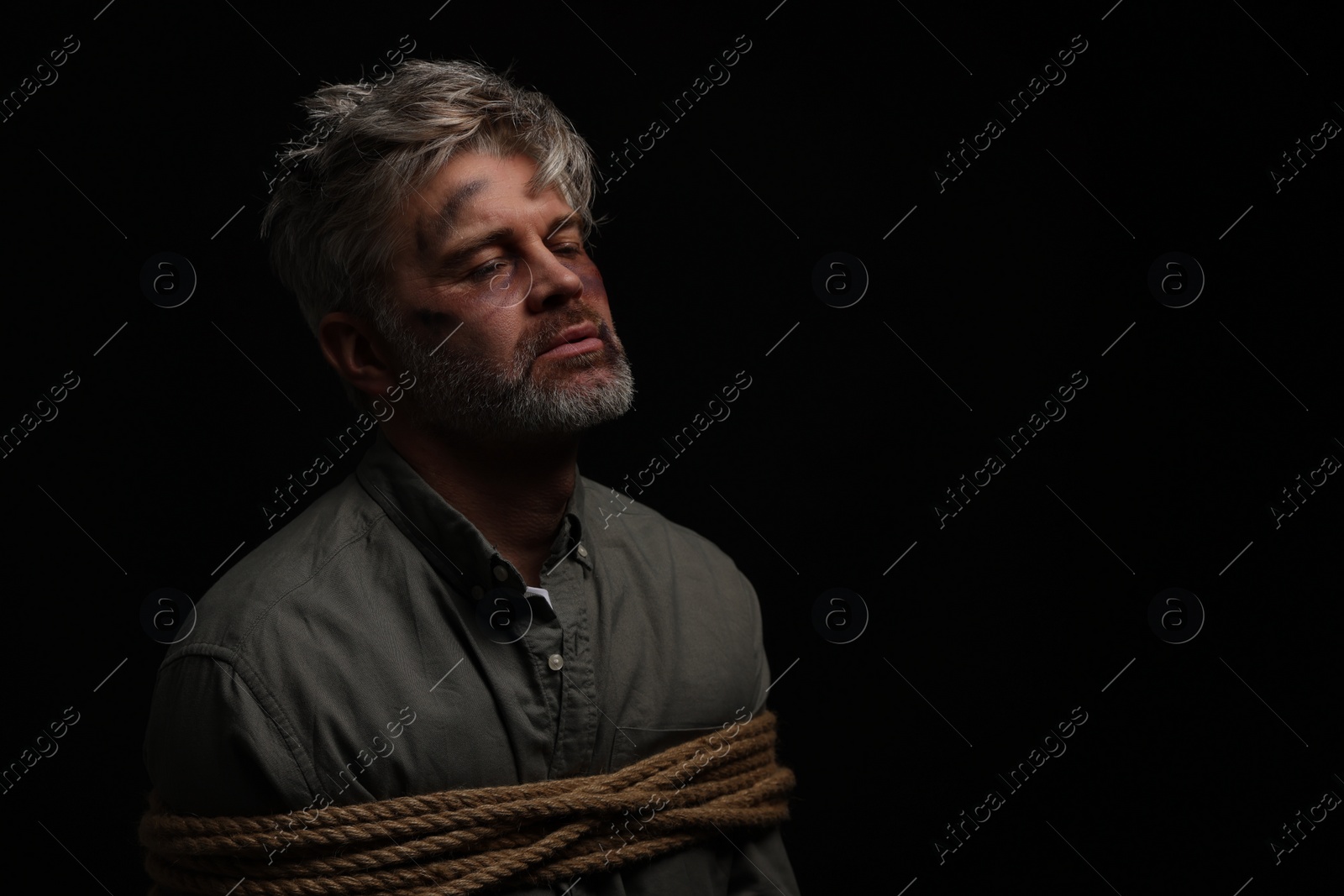 Photo of Beaten man tied with rope on black background, space for text. Hostage taking