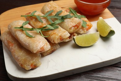 Photo of Tasty fried spring rolls, lime, arugula and sauce on wooden table, closeup