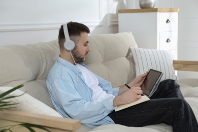 Photo of Young man with headphones using modern tablet for studying on sofa at home. Distance learning