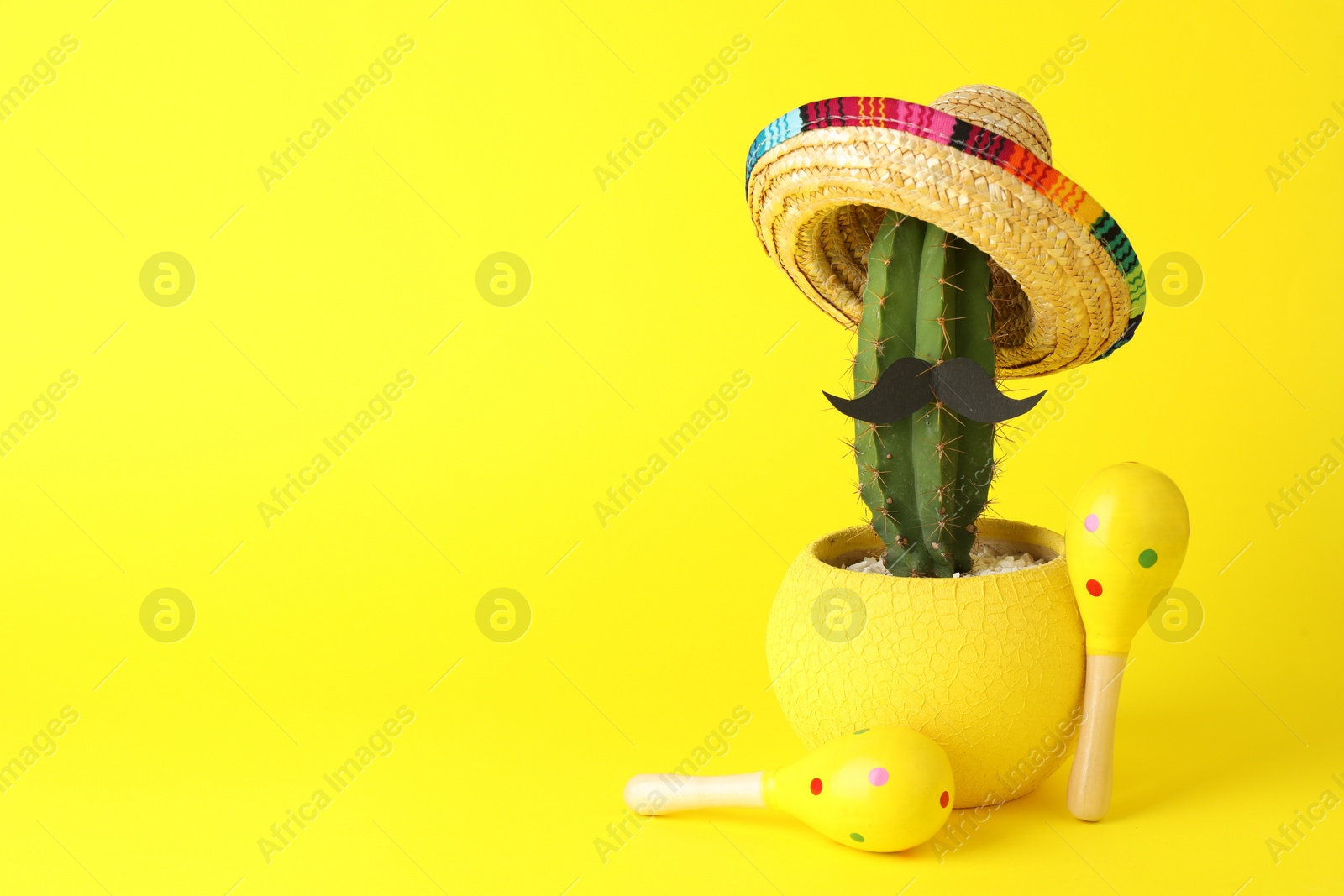 Photo of Cactus with Mexican sombrero hat, fake mustache and maracas on yellow background, space for text