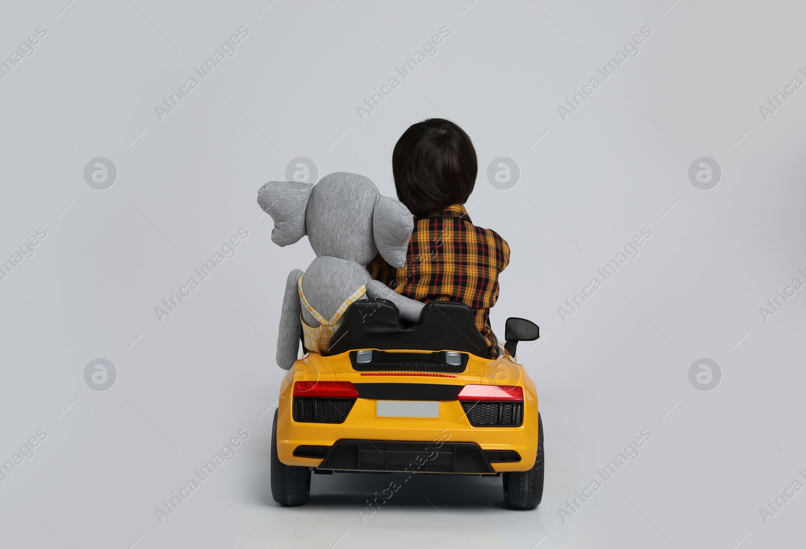 Photo of Cute little boy with toy elephant driving children's car on grey background, back view