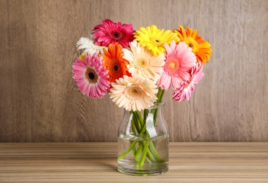 Photo of Bouquet of beautiful bright gerbera flowers in glass vase on table against wooden background