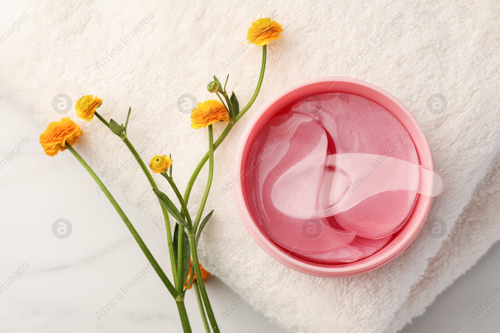 Photo of Jar of under eye patches with spoon, flowers and towel on white marble table, flat lay. Care products