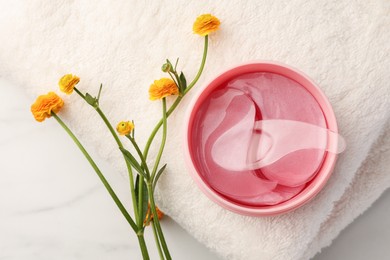 Jar of under eye patches with spoon, flowers and towel on white marble table, flat lay. Care products