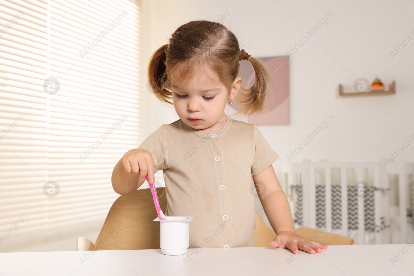 Photo of Cute little child eating tasty yogurt from plastic cup with spoon at white table indoors