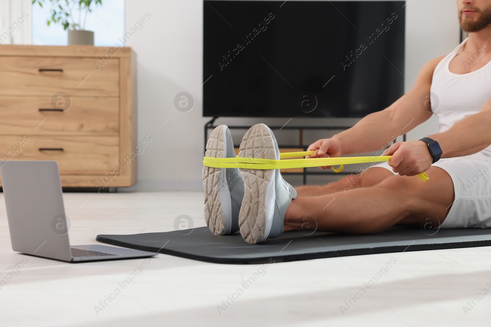 Photo of Athletic man doing exercise with elastic resistance band near laptop on mat at home, closeup
