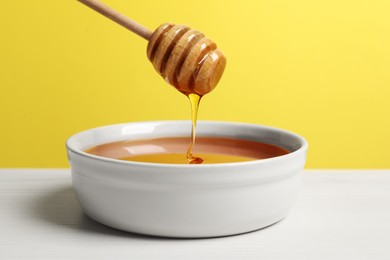 Photo of Pouring tasty honey from dipper into bowl at white wooden table against yellow background, closeup