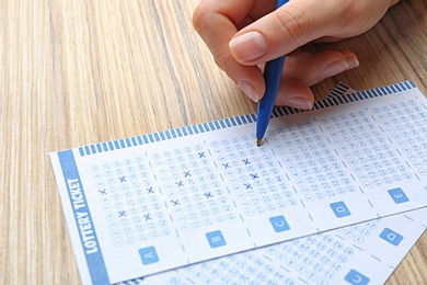 Woman filling out lottery tickets with pen on wooden table, closeup. Space for text