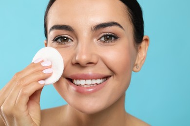 Young woman using cotton pad with micellar water on light blue background, closeup