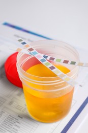 Photo of Container with urine sample for analysis and test strips on white table, closeup