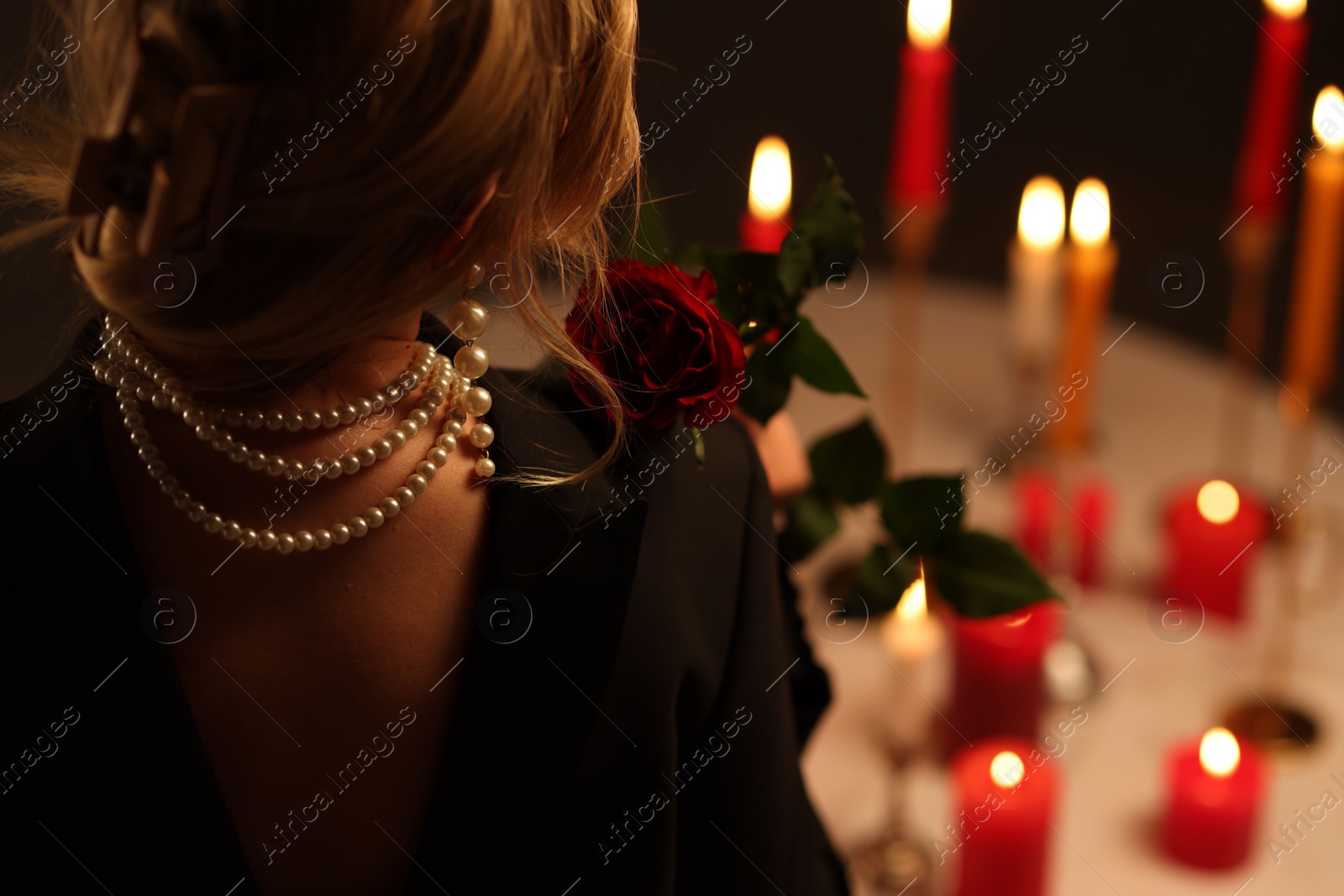 Photo of Woman wearing elegant jewelry near table with burning candles at night, back view