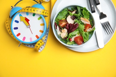 Plate of tasty salad, alarm clock and measuring tape on yellow background, flat lay with space for text. Nutrition regime