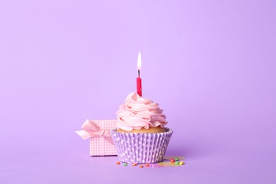 Birthday cupcake with burning candle and gift box on violet background