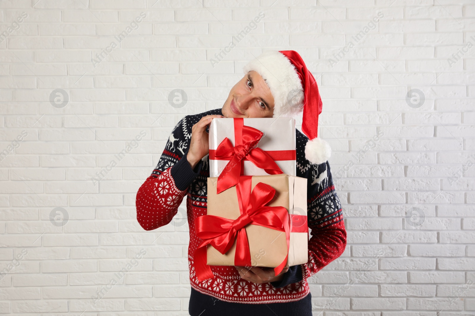 Photo of Happy man in Christmas sweater and Santa hat holding gift boxes near white brick wall