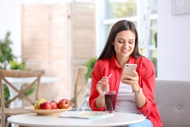 Photo of Young woman using mobile phone while drinking tasty healthy smoothie at table, indoors