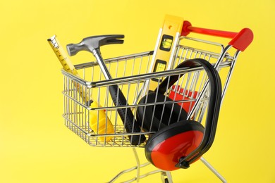 Photo of Small shopping cart with set of construction tools and headphones on yellow background