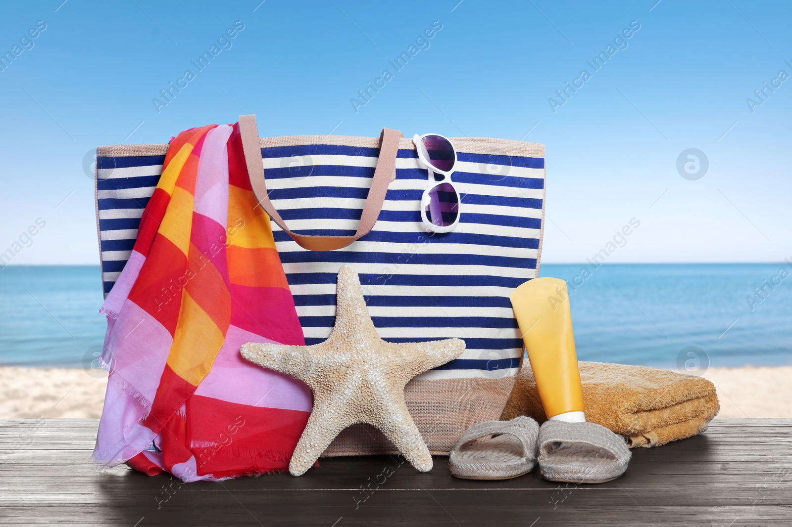 Image of Stylish striped bag and other beach accessories on wooden table near sea